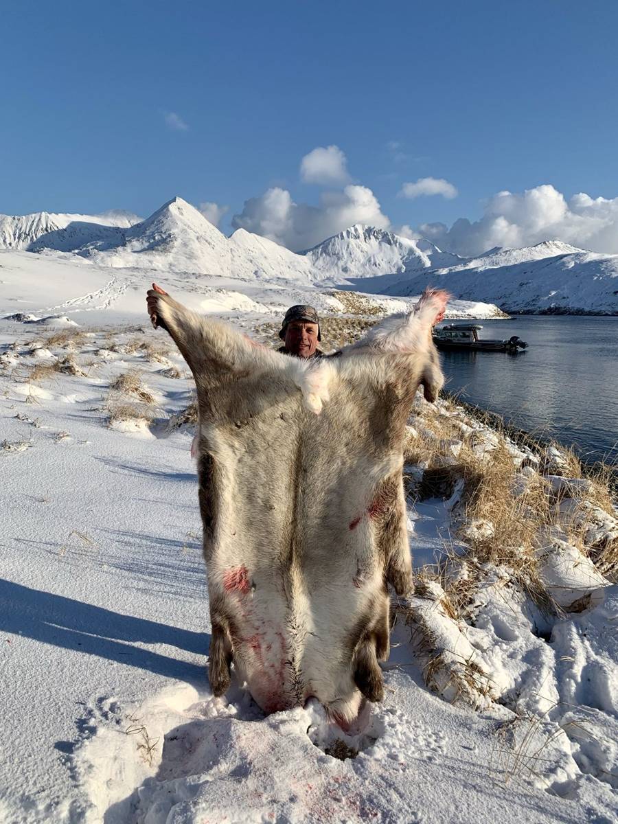 Hunter holding a stretched Caribou hide with snowy Adak mountains and the boat behind