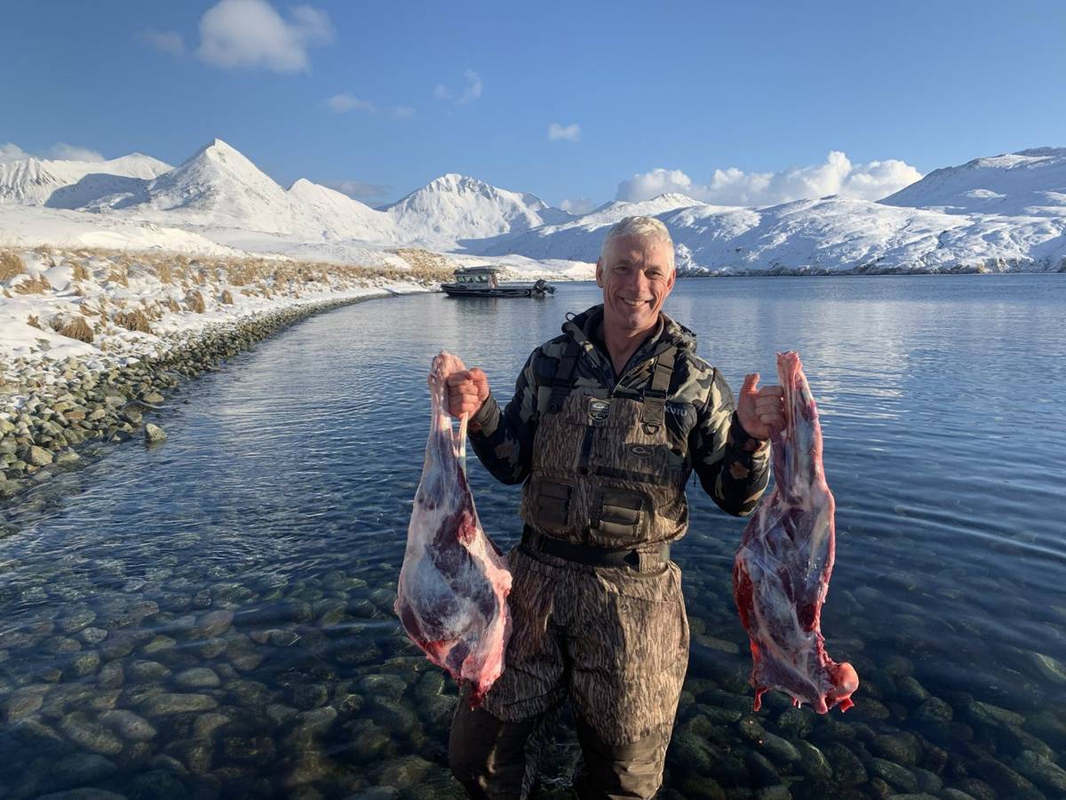 Hunter in the shallows holding two Caribou hindquarters with snowy peaks behind